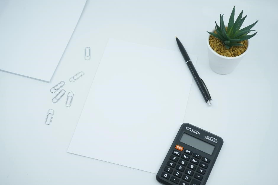 A financial and money themed photo featuring a blank paper on a desk next to a pen and calculator. Crunch the numbers!ure